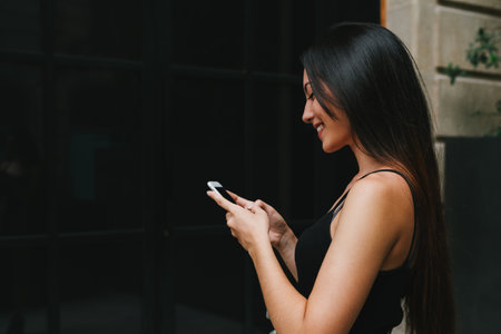 Side view portrait of a beautiful smiling caucasian hipster girl with long dark hair wearing black top surfing web on a mobile phone while standing on a street. Young woman using banking app on mobileの写真素材