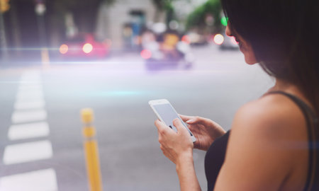 View from the back photo of female checking messages on a mobile phone while standing at the road side. Woman with dark hair using smartphone while standing on blurred road background. Flare light.の写真素材