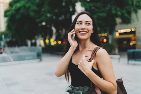 Young brunette caucasian woman with long hair is talking by a smartphone while walking down the street on a blurred urban background. Stylish student girl is chatting with friends by a mobile phone.の写真素材
