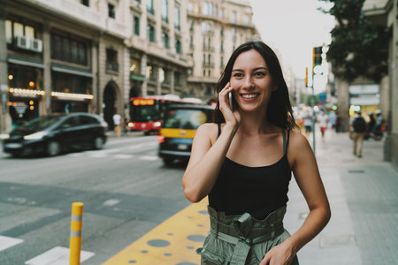 Cheerful hipster girl chatting with a friend on mobile phone while walking the street. Charming student girl wearing stylish clothes calling to a restaurant to reserve a table by a smartphone.の写真素材
