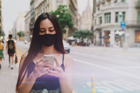 Half length shot of a young woman wearing protective mask chatting with friends via social media by mobile phone. Caucasian female in black mask happy to get email with negative COVID19 test resultsの写真素材