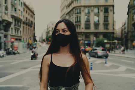 Half length portrait of a beautiful brunette hipster girl with a pensive look wearing black protective mask and looking aside while standing on a blurred urban background during COVID 19 pandemic.の写真素材