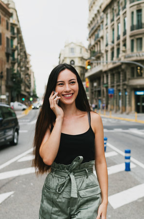 Cheerful caucasian hipster girl standing at the city street and talking on her smartphone. Young attractive woman in trendy outfit having phone conversation while standing on a city road backgroundの写真素材