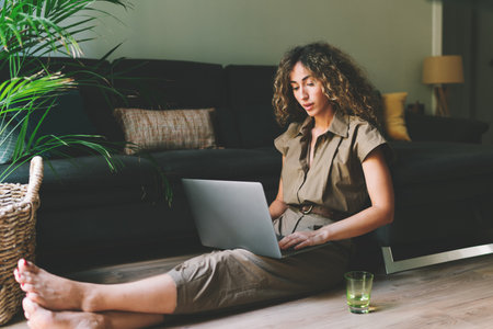 Beautiful female in casual outfit chilling on a floor beside comfortable couch in a living room with laptop on her knees. Web designer browsing laptop while working on a remote project at home.の写真素材