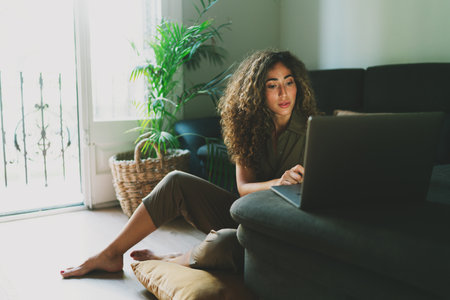 Beautiful hispanic woman working on a laptop computer while sitting on a floor at home during self-isolation at covid19 pandemic period. Beauty blogger writing post using netbook while working at homeの写真素材