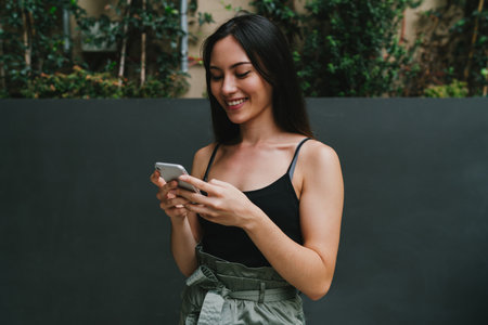 Successful fashion blogger surfing the web on a mobile phone while standing on a blank street wall...