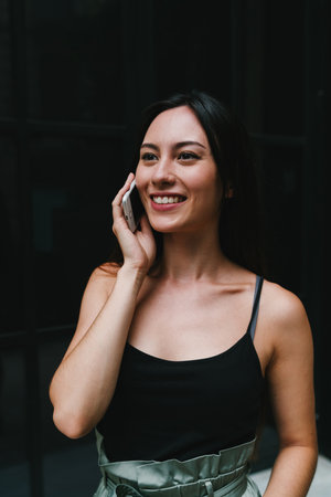 Young business woman happy to receive a call by a mobile phone with good business news. Cheerful student girl talking with friend by a smartphone while standing on a street beside a big dark windowの写真素材