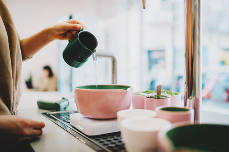 Side view photo of a woman's hand holding a jug with water pouring into the pink bowl with a whipped green matcha tea powder. The process of a japanese green tea making in a coffee shop.の写真素材