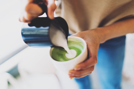 Professional female barista adding whipped milk to a freshly made matcha tea. Woman's hands holding a cup of a matcha tea and a jug with a hot whipped milk. Adding whipped milk to the japanese tea.の写真素材