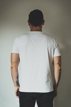 Young hipster man wearing blank white t-shirt with empty space for logo, text or design. mock-up of t-shirt, white wall in the background. Back view. Vertical photoの写真素材