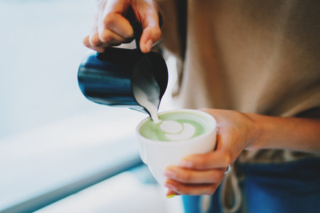 Barista making patterns while making tasty and healthy matcha latte tea. Female's hand pouring hot whipped milk into the cup with freshly made natural matcha tea in a modern asian coffee shop.の写真素材
