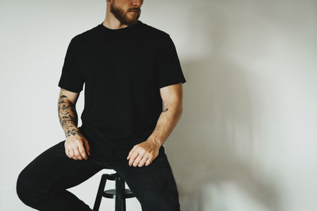 young hipster dressed black t-shirt with empty space for logo, text or design sits on a bar stool against a white wall. mock-up of t-shirt, white wall in the background.の写真素材