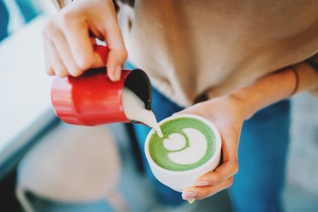 Barista making flower patterns while preparing tasty and healthy matcha latte tea. Woman's hand pouring hot whipped milk into the cup with freshly made natural matcha tea in an asian coffee shop.の写真素材