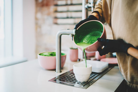 Matcha tea made of natural ingredients by professional barista in asian cafe. Female's hands holding a bowl with a freshly made matcha pouring to the white cup. Tea ceremony in an asian style.の写真素材