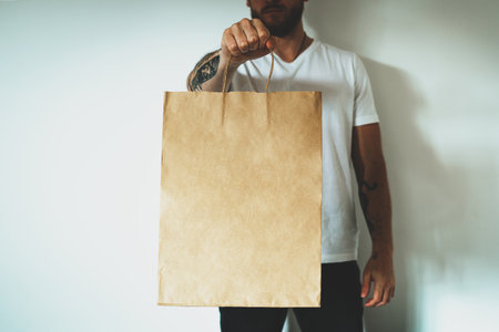 Young hipster man wearing blank white t-shirt and holding craft paper package with empty space for your logo or design, mock-up of shopping bag with handles.の写真素材