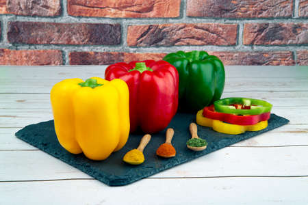 Whole and sliced red, green, yellow peppers and three wooden spoons with spices on black stone board and brick wall,の写真素材