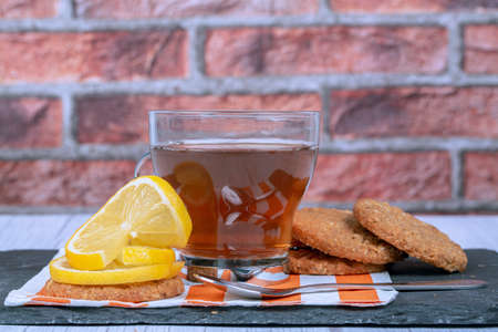 Oatmeal cookies, cup of tea, teaspoon, lemon slices and, on stone plate and striped napkin with a brick backgroundの写真素材