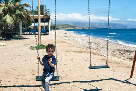 Sweet little boy on a swing right on the beach, looking straight ahead with curly hair on a sunny day.の写真素材