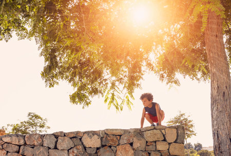 a little boy enjoys climbing a stone wall, under a tree through which the sun's rays enter at sunset.の写真素材