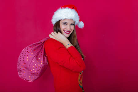 beautiful smiling woman with Santa Claus hat and Christmas reindeer sweater, carrying sack of presents on her backの写真素材