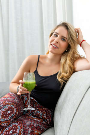 young blonde woman sitting on the sofa drinking a vegetable smoothieの写真素材