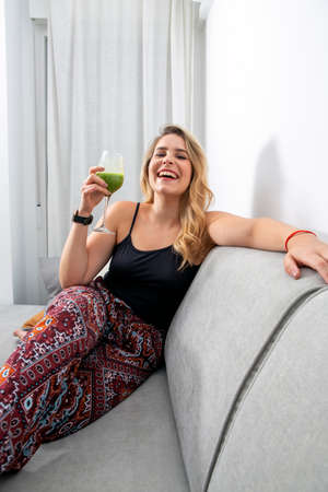 young blonde woman sitting on the sofa drinking a vegetable smoothie
