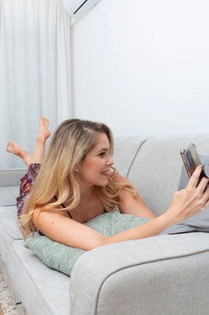 young blonde woman taking photos with her smart phone on the sofa in her living roomの写真素材