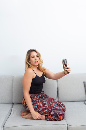 pretty blonde woman taking photos with her smart-phone on the sofa in her living roomの写真素材