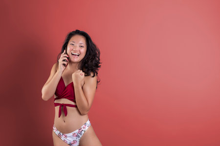 young asian ethnic woman in bikini talking on mobile phone, on plain backgroundの写真素材