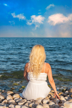 woman in white dress, seen from behind, sitting on a pebble beach shore, looking at the sea.の写真素材