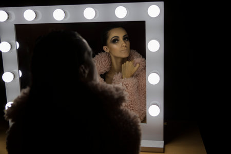 Attractive girl looking at her beauty in light mirror with pink coatの写真素材