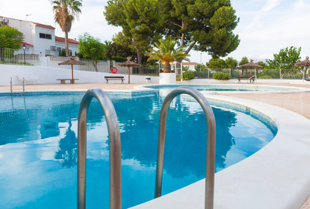 Residential swimming pool with ladder access, surrounded by greenery and adjacent modern homes.