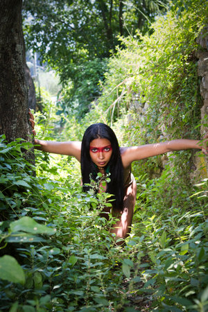 An indigenous Brazilian woman with traditional face paint exploring a dense jungle, surrounded by lush green vegetation.の写真素材