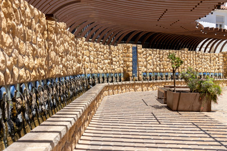 A long stone wall fountain under an arched wooden pergola in an outdoor public space with ample sunlight.の写真素材