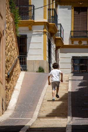 A young boy in casual clothing climbs stone stairs on a sunny, narrow street, surrounded by traditional Mediterranean architecture.の写真素材