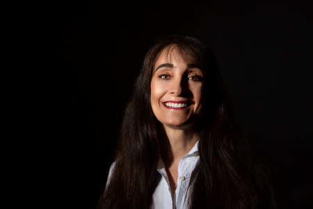 A portrait of a smiling woman with long dark hair, wearing a white shirt, captured with dramatic lighting against a dark background.の写真素材