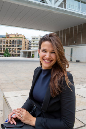 Confident businesswoman smiling brightly while sitting outdoors in an urban environment, with modern architecture and buildings in the background.の写真素材