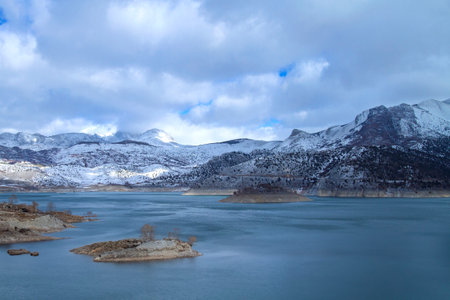 Scenic winter landscape of a snow-covered mountain range and calm lake, with dramatic clouds overhead, creating a serene atmosphere.の写真素材