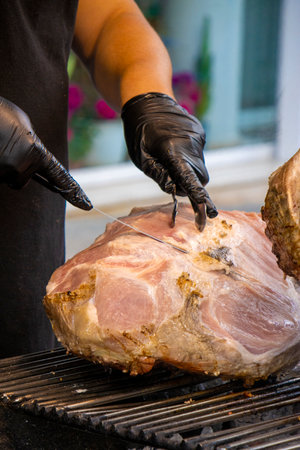 Close-up of a person slicing freshly roasted ham with a knife, wearing black gloves at a traditional...