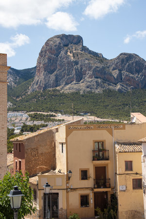 Traditional stone houses with terracotta roofs sit beneath a towering rocky mountain, with lush green forested slopes and a bright blue sky with scattered clouds aboveの写真素材