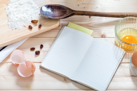 Old  blank recipe cookery book on wooden background with copy space, egg yoke, wooden, spoon egg shell raisins, flour, board, and yellow post it paper の写真素材