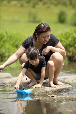 Young Asian mother and daughter playing paper boat by riverの写真素材