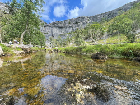 Malham, A tranquil scene unfolds with a clear river reflecting a rugged cliff face, framed by lush...
