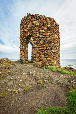 A stone tower stands on a cliff overlooking the ocean, bathed in sunlight under a cloudy sky....