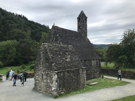An ancient stone church Glendalough with a round tower, surrounded by lush greenery and a cloudy sky in Ireland.の写真素材