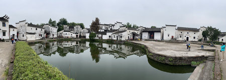 A tranquil village scene in China, featuring traditional white houses reflected in a serene pond under an overcast sky.の写真素材