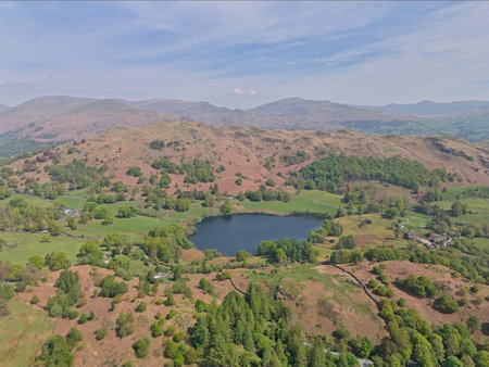An aerial view captures a scenic lake surrounded by lush green hills and distant mountains under a partly cloudy sky.の写真素材