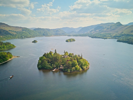 Aerial view of Derwentwater Lake with green islands and surrounding mountains under a clear blue sky. A boat is sailing on the water.の写真素材
