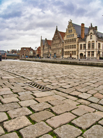 Cobblestone View of Ghent's Guild Houses - The historic guild houses of Ghent line the canal under a cloudy sky, with a focus on the wide, textured cobblestone street and a storm drain in the foreground.の写真素材
