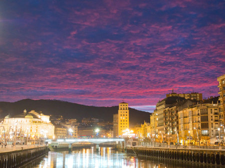 Sunrise colors over Bilbao cityscape. - The NerviÃ³n River and city skyline of Bilbao at dawn, with buildings and streetlights reflected on the water under a pink and purple sky.の写真素材